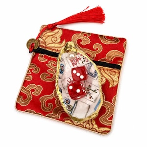 May include: A square red fabric pouch with gold cloud patterns, a black zipper, and a red tassel with a brass coin. Resting on top is an oyster shell with a gold-painted edge, containing two red dice with white pips and a single white pearl.
