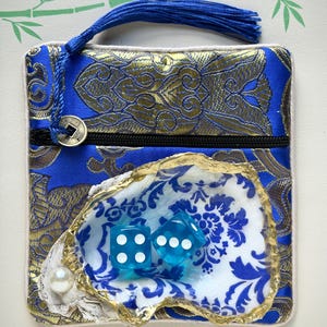 K&ouml;nnte beinhalten: Ein quadratischer Beutel aus blau-goldenem Brokat mit schwarzem Rei&szlig;verschluss, blauer Quaste und M&uuml;nzanh&auml;nger. Auf der Vorderseite befindet sich eine Austernschale mit goldfarbenem Rand, einer wei&szlig;en Perle und zwei blauen W&uuml;rfeln.