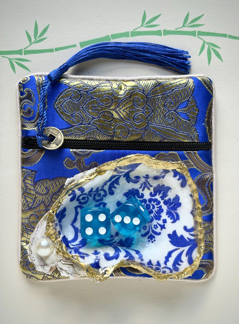 K&ouml;nnte beinhalten: Ein quadratischer Beutel aus blau-goldenem Brokat mit schwarzem Rei&szlig;verschluss, blauer Quaste und M&uuml;nzanh&auml;nger. Auf der Vorderseite befindet sich eine Austernschale mit goldfarbenem Rand, einer wei&szlig;en Perle und zwei blauen W&uuml;rfeln.