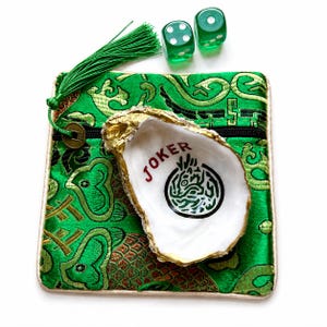 May include: A green fabric pouch with a tassel and a gold-toned coin charm, two translucent green dice, and a white oyster shell with gold-painted edges. The shell features the word JOKER in red text and a circular black and green graphic design.