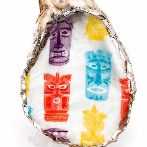 May include: A decorative oyster shell featuring a white interior with a repeating pattern of colorful tiki masks in purple, yellow, red, and blue. The shell edge is finished with a metallic silver trim, and a single white pearl is set at the top.