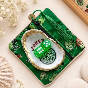 May include: A square green fabric pouch with floral embroidery, a black zipper, and a green tassel. Resting on the pouch is a white oyster shell with a gold-painted rim, containing two translucent green dice and the red text JOKER.