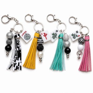 May include: Four keychains featuring silver-tone lobster clasps and rings. Each includes a mahjong tile, a leather-like tassel, and a strand of decorative beads in various colors, patterns, and textures including black, white, silver, yellow, teal, and pink.