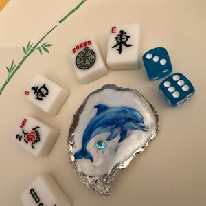 May include: A white oyster shell with a painted blue dolphin and a small blue rhinestone, surrounded by five white Mahjong tiles with black and red markings and two translucent blue dice with white pips arranged on a light surface.