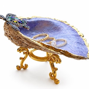 May include: A decorative oyster shell dish with a purple interior and gold-painted edges, resting on a gold-toned metal stand. A jeweled turtle figurine with blue and iridescent stones is attached to the shell, which holds three silver rings.