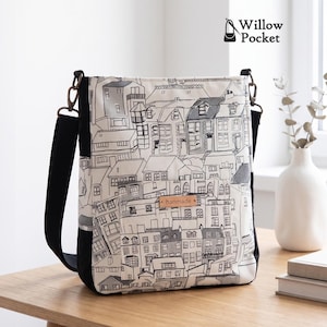 Puede incluir: Un bolso bandolera con un estampado de dibujo lineal en blanco y negro de un paisaje urbano con casas. El bolso tiene paneles laterales negros, una correa ajustable negra con herrajes metálicos y una pequeña etiqueta beige con el texto hanmade.