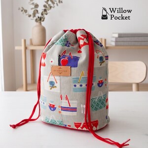 Puede incluir: Una bolsa de cordón gris claro con un patrón colorido de varios bolsos y artículos de cocina. La bolsa tiene cordones de color rojo brillante y una etiqueta rectangular de color canela en la parte frontal con la palabra HANDMADE impresa.