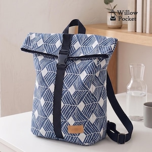 May include: A roll-top backpack featuring a blue and white geometric leaf pattern. The bag includes a black vertical strap with a plastic buckle closure, adjustable black shoulder straps, and a small tan leather-like label labeled handmade.