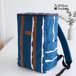May include: A rectangular backpack featuring a blue fabric with white vertical brushstroke patterns. It includes two brown vertical straps, a brown rectangular label with the word handmade, and a brown zipper closure. The logo Willow Pocket is visible.
