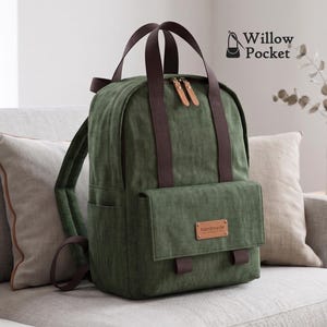 Puede incluir: Una mochila de color verde oliva oscuro con correas y asas de tela marrón oscuro. La bolsa cuenta con un bolsillo frontal con un parche rectangular de cuero sintético color canela con la palabra handmade y el logo Willow Pocket.