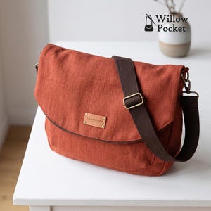 May include: A rust-colored messenger bag with a textured fabric finish, featuring a dark brown adjustable webbing strap, antique brass hardware, and a rectangular leather-like patch embossed with the word handmade on the front flap.