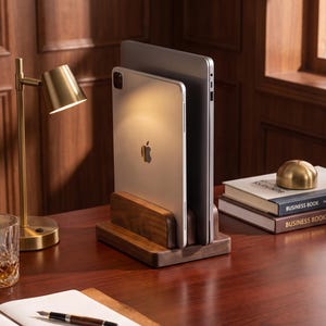 May include: A dark brown wooden vertical stand holding a silver tablet and a silver laptop. The stand features two parallel slots designed to keep electronic devices upright. The wood has a smooth, polished finish with visible natural grain patterns.