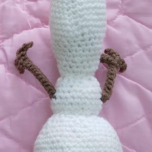 May include: A white crocheted snowman toy with brown crocheted arms and a brown crocheted top. The snowman is lying on a pink surface.