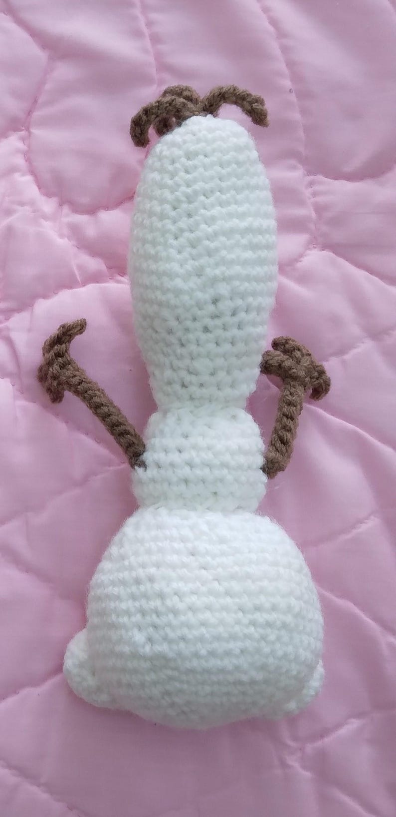 May include: A white crocheted snowman toy with brown crocheted arms and a brown crocheted top. The snowman is lying on a pink surface.