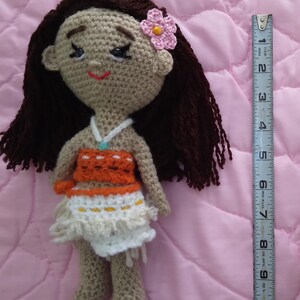K&ouml;nnte beinhalten: H&auml;kelpuppe mit braunen Haaren, einer rosa Blume und einem braunen, orangen und wei&szlig;en Outfit. Die Puppe ist etwa 25 cm gro&szlig;.
