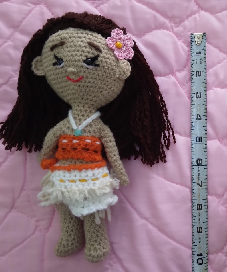 K&ouml;nnte beinhalten: H&auml;kelpuppe mit braunen Haaren, einer rosa Blume und einem braunen, orangen und wei&szlig;en Outfit. Die Puppe ist etwa 25 cm gro&szlig;.