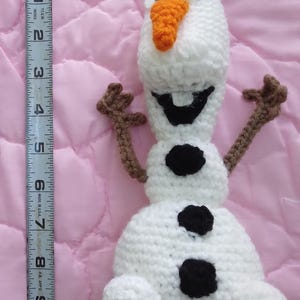 May include: A white crocheted snowman with brown arms and a carrot nose. The snowman has black eyes and a black mouth. The snowman is about 7 inches tall.