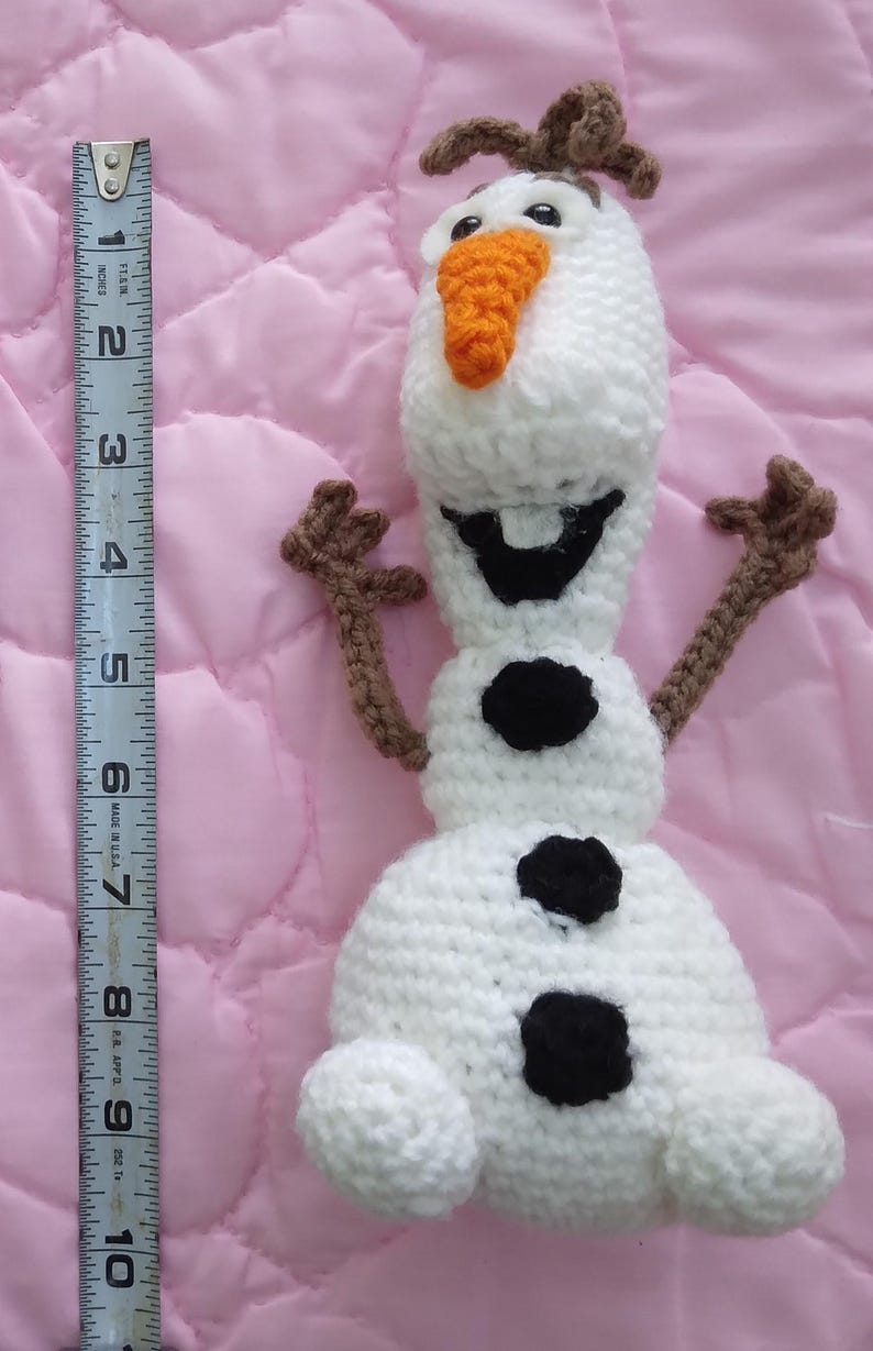 May include: A white crocheted snowman with brown arms and a carrot nose. The snowman has black eyes and a black mouth. The snowman is about 7 inches tall.