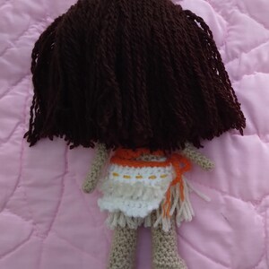 K&ouml;nnte beinhalten: Eine geh&auml;kelte Puppe mit braunem Garnhaar, die ein wei&szlig;es und orangefarbenes Kleid mit Fransen tr&auml;gt. Die Puppe steht auf einem rosa gesteppten Stoff.