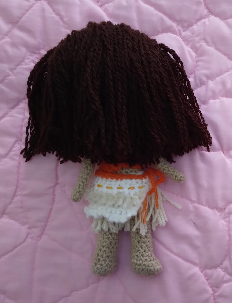 K&ouml;nnte beinhalten: Eine geh&auml;kelte Puppe mit braunem Garnhaar, die ein wei&szlig;es und orangefarbenes Kleid mit Fransen tr&auml;gt. Die Puppe steht auf einem rosa gesteppten Stoff.