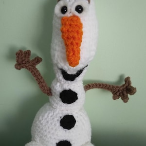 May include: A crocheted Olaf snowman toy with brown arms, a white body, black buttons, and an orange carrot nose.
