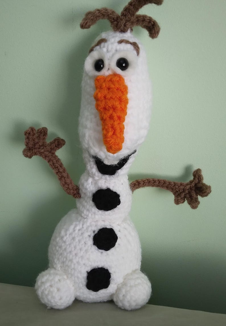 May include: A crocheted Olaf snowman toy with brown arms, a white body, black buttons, and an orange carrot nose.