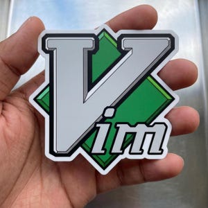 May include: A die-cut sticker featuring the Vim text logo in a bold, silver-grey font with a thick black outline. The design is layered over a green diamond-shaped background with a subtle three-dimensional beveled edge effect.