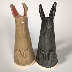May include: Two ceramic rabbit figurines with tall, upright ears and cylindrical bodies. One rabbit is light brown with pink-lined ears, while the other is dark charcoal gray. Both feature small, simple facial details and two raised circular feet.