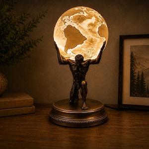 May include: A bronze-toned table lamp featuring a kneeling figure supporting a glowing, spherical globe. The globe displays a detailed map of the Earth with illuminated continents and dark oceans, resting on a circular base with a decorative edge.