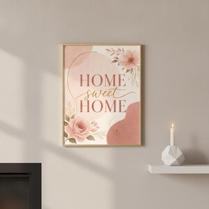May include: A rectangular wall art print in a gold frame featuring the text "HOME sweet HOME" in a mix of serif and script fonts. The design includes soft pink and terracotta abstract shapes, delicate floral illustrations, and gold line accents.