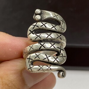Può includere: Un anello in metallo color argento a forma di serpente avvolto. La superficie del serpente presenta un motivo ripetitivo di piccoli segni a X incisi lungo il corpo. La testa del serpente è arrotondata con due piccole rientranze circolari che rappresentano gli occhi.