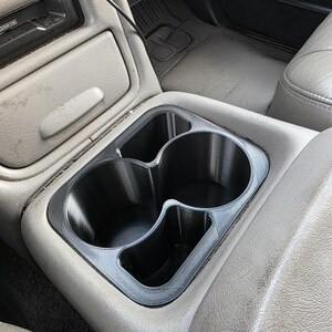 May include: A black plastic cup holder insert with two circular openings and two smaller rectangular compartments, designed to fit into the center console of a vehicle. The insert features a smooth, matte finish and is positioned within a gray interior.