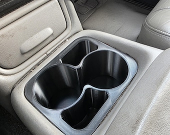 May include: A black plastic automotive cup holder insert featuring two large circular openings and two smaller rectangular compartments, designed to fit into the centre console of a vehicle with a light grey interior finish.