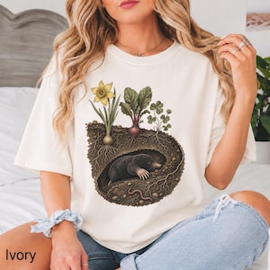 May include: An ivory t-shirt featuring a graphic illustration of a mole inside an underground burrow. Above the burrow, a yellow daffodil, a beet with green leaves, and a clover plant are visible. The soil shows roots and a small pink earthworm.