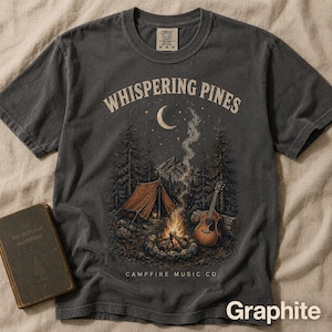 May include: A charcoal grey short-sleeve t-shirt featuring a graphic of a campsite with a tent, a campfire, and an acoustic guitar. The text reads "WHISPERING PINES" above the scene and "CAMPFIRE MUSIC CO." below. The shirt has a crew neck.