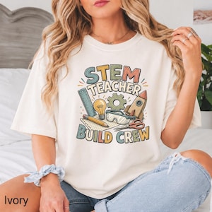 May include: An ivory t-shirt featuring a colorful graphic with the text STEM TEACHER BUILD CREW. The design includes illustrations of a ruler, a lightbulb, a gear, a rocket, safety goggles, a pencil, and scissors in muted tones of blue, orange, and green.
