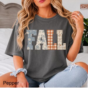 May include: A charcoal gray short-sleeve t-shirt featuring the word FALL printed in large block letters across the chest. Each letter has a distinct pattern: blue floral, cream floral, orange and white gingham, and a cream plaid with small flowers.