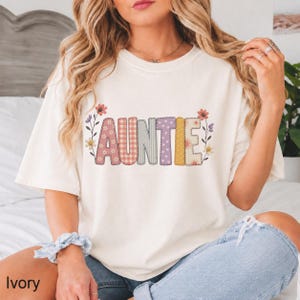 May include: An ivory t-shirt featuring the word AUNTIE in large, blocky letters. Each letter has a unique pattern, including floral, gingham, striped, and polka dot designs in muted tones. Small floral illustrations frame the text on both sides.