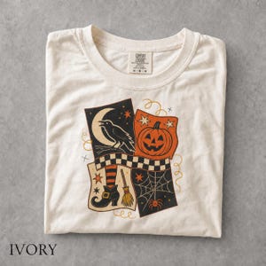 May include: An ivory t-shirt featuring a graphic print of a black raven on a crescent moon, an orange jack-o'-lantern, a witch's boot with striped socks, a broom, and a spider web. The shirt has a crew neck and a Comfort Colors brand label.