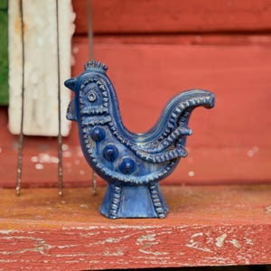 Könnte beinhalten: Eine blaue Keramik-Hahnfigur mit strukturierter Oberfläche, erhabenen kreisförmigen Details, einem Kamm und einem gefächerten Schwanz. Die Glasur weist Variationen in der Farbintensität auf, die die geformten Muster an Hals und Flügeln betonen.