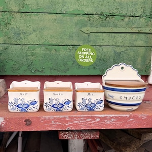 Può includere: Un set di quattro contenitori da cucina in ceramica bianca con motivi floreali blu e coperchi in legno. Tre contenitori rettangolari più piccoli riportano le scritte Salt, Socker e Mjol. Un contenitore rotondo da parete riporta la scritta Mjol.