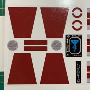 May include: A set of red and white stickers for a model spaceship. The stickers feature a variety of designs, including a control panel, a spaceship window, and a spaceship wing.