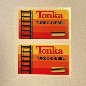 May include: Two Tonka Turbo-Diesel toy truck stickers. Each sticker features a yellow, orange, and red background with a black ladder and a black and yellow wood plank. The text "Tonka Turbo-Diesel" is printed in black.