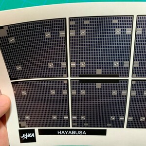 Op de afbeelding: Een close-up van een wit vel papier met een gedrukt rasterpatroon. Het raster is gemaakt van kleine vierkanten en er lopen verschillende witte lijnen over het raster. De tekst "HAYABUSA" is onderaan het vel gedrukt.