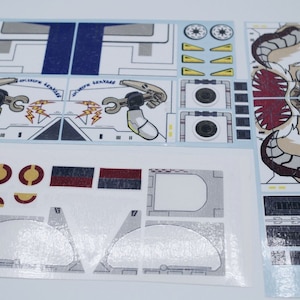 May include: A sheet of stickers for a Lego Star Wars set. The stickers feature a white and gray spaceship with a blue stripe, a white and gray creature with red eyes and teeth, and a white and gray robot arm with a yellow lightning bolt.