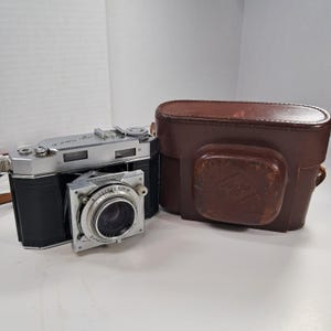 May include: A silver and black folding camera with a lens labeled Synchro, positioned next to a brown leather carrying case. The camera body features metallic accents and black textured grips, while the case has a visible embossed logo.