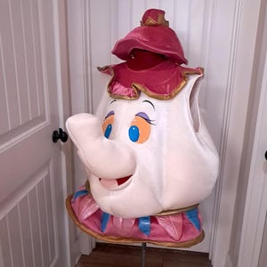 May include: A character costume shaped like a teapot, featuring a white rounded body, a protruding spout, and a pink lid with gold trim. The base is decorated with pink and blue petal-like accents. The face has large blue eyes and a smiling mouth.