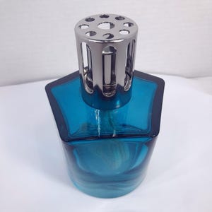 May include: A translucent teal glass fragrance lamp with a pentagonal base and a rounded bottom. The top features a polished silver-colored metal crown cap with circular cutouts, designed to sit securely over the neck of the glass vessel.