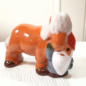 May include: A ceramic figurine featuring a glossy orange horse with a white mane and tail, standing beside a small gnome with a red hat, a long white beard, and a blue coat. The horse has dark brown hooves and the gnome has dark green accents.