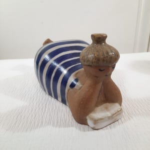 Może przedstawiać: Ceramiczna figurka przedstawiająca osobę leżącą na brzuchu i czytającą książkę. Postać ma teksturowaną brązową głowę i ramiona oraz korpus pomalowany w poziome granatowo-białe paski. Książka jest biała, a postać ma zamknięte oczy.